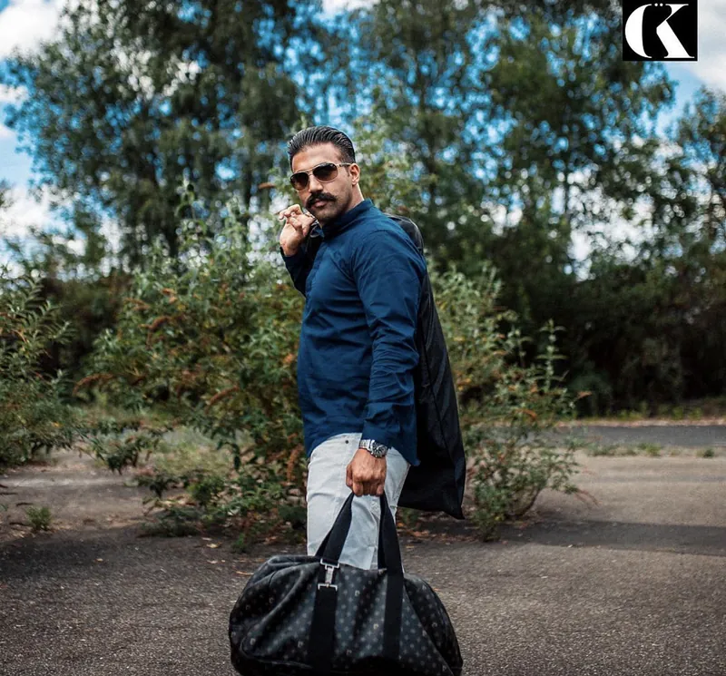 Ömer Kaya mit Sonnenbrille und Reisetasche, das Telefon am Ohr, zwischen Sträuchern in Wiesbaden