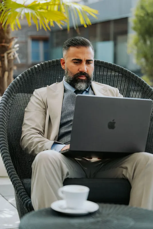 Ömer Kaya im beigen Anzug mit Laptop in einem Korbsessel, davor eine Kaffeetasse